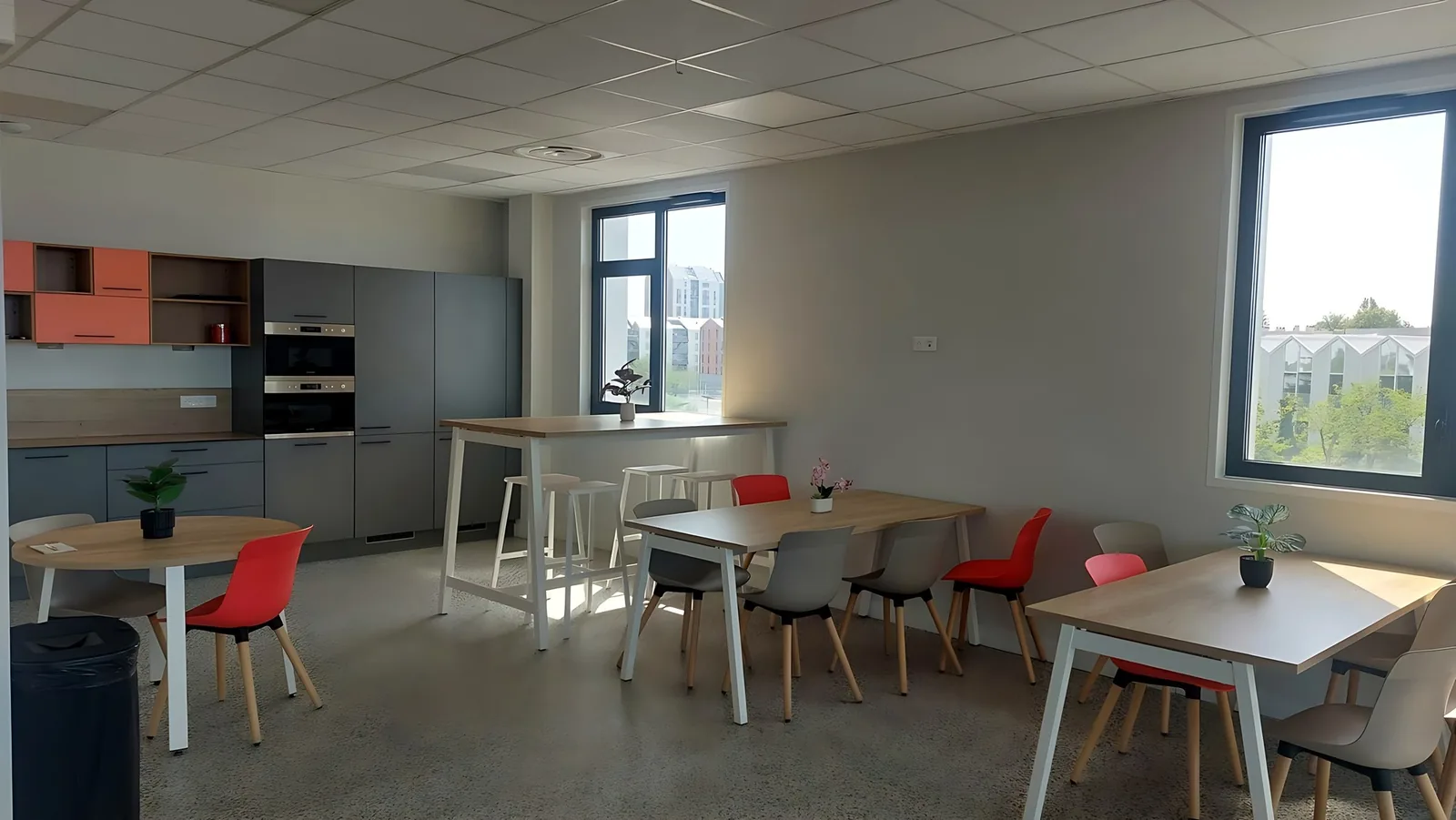 Salle de restauration du campus : cuisine grise, tables de bois clair et chaises corail devant de grandes fenêtres